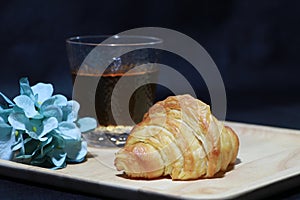The croissant placed on the square wooden tray with tea in a glass and blue flowers on dark background
