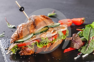 Croissant sandwich with fillet grilled chicken, fresh vegetables, cheese and greens on black shale board over black stone