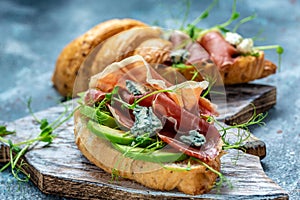 Croissant sandwich with jamon ham serrano paleta iberica, blue cheese, avocado, microgin on blue background. Food recipe