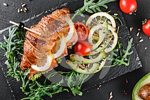 Croissant sandwich with red fish, avocado, fresh vegetables and arugula on black shale board over black stone background