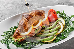 Croissant sandwich with red fish, avocado, fresh vegetables and arugula on black shale board over black stone background