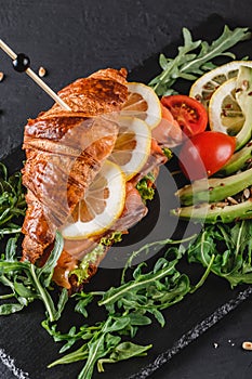 Croissant sandwich with red fish, avocado, fresh vegetables and arugula on black shale board over black stone background