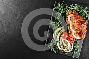 Croissant sandwich with red fish, avocado, fresh vegetables and arugula on black shale board over black stone background