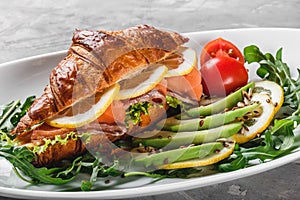 Croissant sandwich with red fish, avocado, fresh vegetables and arugula on black shale board over black stone background