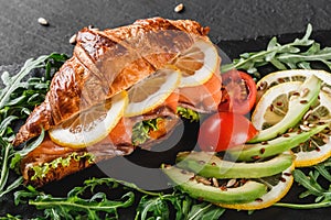 Croissant sandwich with red fish, avocado, fresh vegetables and arugula on black shale board over black stone background