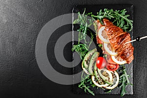 Croissant sandwich with red fish, avocado, fresh vegetables and arugula on black shale board over black stone background