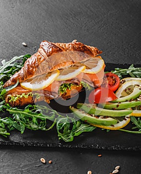 Croissant sandwich with red fish, avocado, fresh vegetables and arugula on black shale board over black stone background