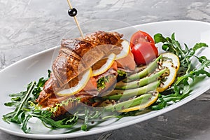 Croissant sandwich with red fish, avocado, fresh vegetables and arugula on black shale board over black stone background