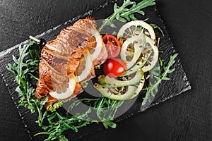 Croissant sandwich with red fish, avocado, fresh vegetables and arugula on black shale board over black stone background