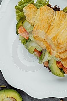 Croissant sandwich with red fish, avocado, fresh vegetables and arugula on black shale board over black stone background
