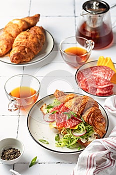 Croissant sandwich with salami sausage, cheese and cucumber in a plate with cup of tea for tasty breakfast on white tile