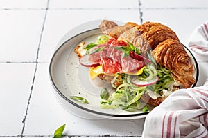 Croissant sandwich with salami sausage, cheese and cucumber in a plate for tasty breakfast on white tile table, text