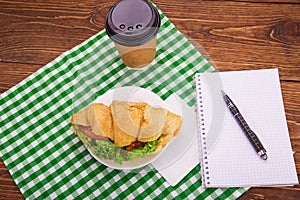 Croissant sandwich on a saucer. Notepad and pen on a wooden table.