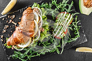 Croissant sandwich with tuna, avocado, fresh arugula and greens on black shale board over black stone background