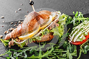 Croissant sandwich with tuna, avocado, fresh arugula and greens on black shale board over black stone background