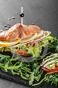 Croissant sandwich with tuna, avocado, fresh arugula and greens on black shale board over black stone background. Healthy food