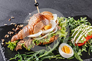 Croissant sandwich with tuna, avocado, fresh arugula and greens on black shale board over black stone background. Healthy food