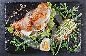 Croissant sandwich with tuna, eggs, avocado, fresh arugula and greens on black shale board over black stone background