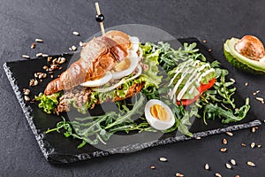 Croissant sandwich with tuna, eggs, avocado, fresh arugula and greens on black shale board over black stone background