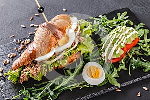 Croissant sandwich with tuna, eggs, avocado, fresh arugula and greens on black shale board over black stone background