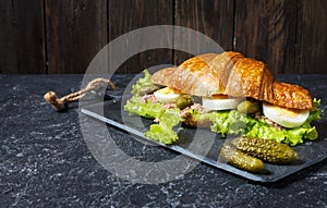 Croissant sandwich with tuna, hard boiled egg, salad and cucumber on stone table