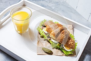 Croissant sandwich with tuna, hard boiled egg, salad and cucumber on white tray. Glass of orange juice. Breakfast
