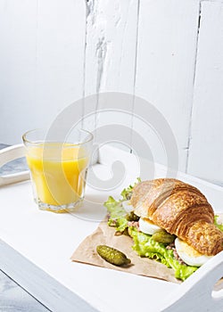 Croissant sandwich with tuna, hard boiled egg, salad and cucumber on white tray. Glass of orange juice. Breakfast concept