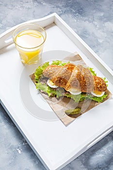 Croissant sandwich with tuna, hard boiled egg, salad and cucumber on white tray. Glass of orange juice. Breakfast