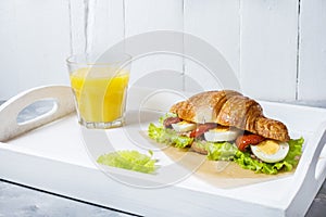 Croissant sandwich with tuna, hard boiled egg, salad and sun-dried tomatoes on white tray. Glass of orange juice. Breakfast concep