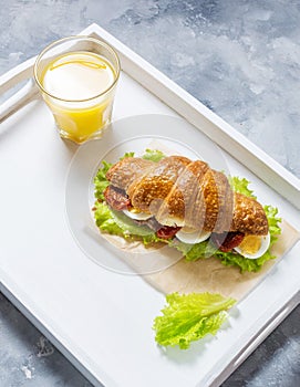 Croissant sandwich with tuna, hard boiled egg, salad and sun-dried tomatoes on white tray. Glass of orange juice.