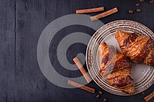 Croissants on a ceramic plate, cinnamon sticks and coffee beans on a black wooden table. Top view with space for text.