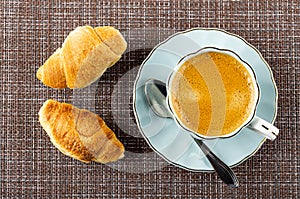 Croissants, cup with black coffee, spoon on saucer on mat. Top view