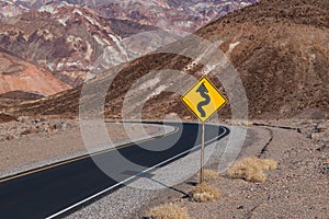A crooked road sign along a highway curving through a barren and rugged desert landscape
