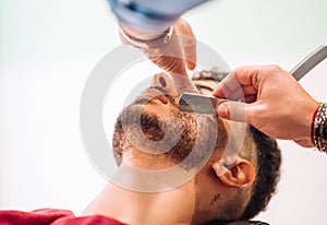 Crop barber cutting beard of client with razor