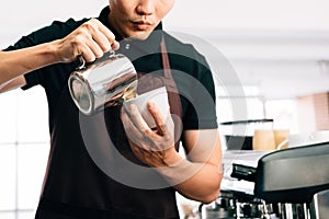 Crop image of a young barista wearing an apron pouring hot milk into hot espresso black coffee for making Latte Art