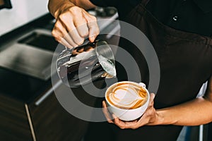 Crop image of a young male barista pouring hot milk into hot espresso black coffee for making Latte Art