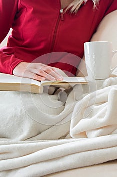 Crop view of young adult woman is relaxing on comfortable sofa with book and hot beverage