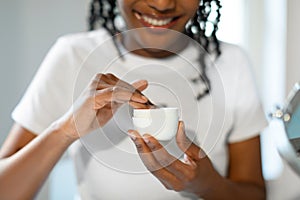 Cropped of black woman using face cream at home, testing newest organic beauty product, holding plastic jar, closeup