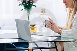 Cropped blond smiling woman cup coffee, light morning white laptop glass desk