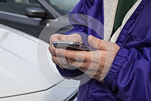 Cropped close-up of man in purple jacket holding a phone in front of parked cars - selective focus