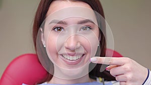 Cropped close up photo of young smiling girl pointing index finger on healthy toothy smile sitting at dental office chair