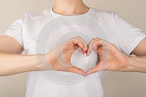 Cropped closeup photo of girl in white t-shirt making heart with fingers near left side of chest on  grey background