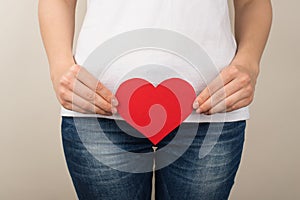 Cropped closeup photo of woman in white t-shirt and jeans holding red paper heart near crotch on  grey background