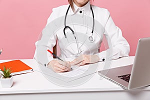 Cropped female doctor siting at desk working filling out medical documents in hospital isolated on pastel pink wall