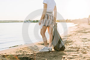 Cropped Female volunteer with bag and gloves picking up trash, a plastic bottles, clean up beach with a sea. Woman