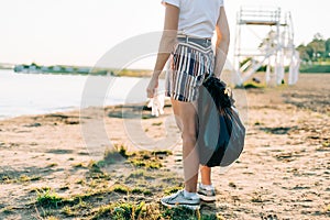 Cropped Female volunteer with bag and gloves picking up trash, a plastic bottles, clean up beach with a sea. Woman