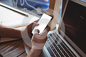 Cropped hand of man using mobile phone over laptop in cafe