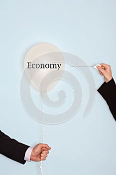 Cropped hands holding needle and popping balloon against light blue background with the text saying Economy