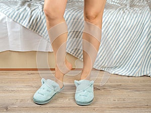 Cropped image of adult woman slender legs putting on soft home slippers, standing by the bed. Woman feet in blue cozy slippers on