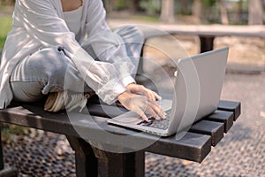 A cropped image of an Asian woman sits on a bench in a public park and uses her laptop computer